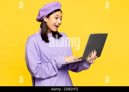 donna asiatica felice in beret utilizzando un computer portatile isolato sul giallo Foto Stock