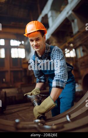 Lavoratore maschile in uniforme e casco rimuove la bilancia da pezzi di metallo in fabbrica. Industria metallurgica, produzione industriale di prodotti siderurgici Foto Stock