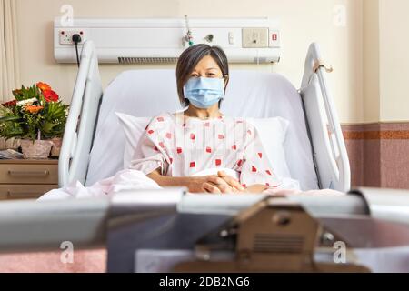 Donna asiatica allegra con maschera facciale a letto in ospedale reparto alla ricerca di cure mediche Foto Stock