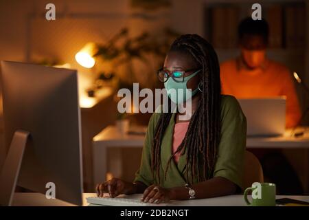 Ritratto di giovane afroamericana indossando maschera e occhiali mentre si lavora con il PC in ufficio di notte, copia spazio Foto Stock