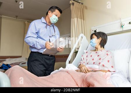 Fiducioso medico asiatico con maschera facciale che si espiano a allegra paziente femminile in condizioni mediche sdraiato a letto in ospedale. Foto Stock