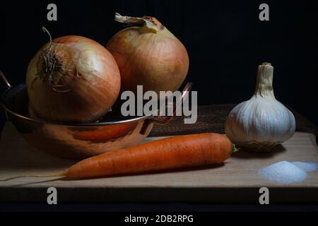 Immagine di cipolle, carote e aglio con un pizzico di sale su tagliere di legno con sfondo nero Foto Stock