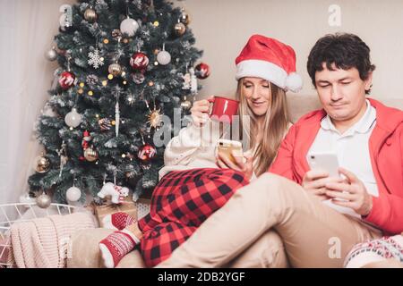 Ritratto di bella coppia marito e moglie in cappello rosso Santa con i loro telefoni cellulari godersi le vacanze invernali di Natale a casa. Foto Stock