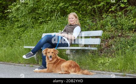 Donna si siede sul banco del parco, cane di razza mista che giace di fronte ad esso. Non per guide animali fino al 9/2022 Foto Stock