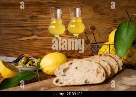 Limoncello italiano con olive e pane fresco su fondo rustico in legno. Liquore tradizionale fatto in casa al limone con agrumi freschi. Primo piano con Short Foto Stock