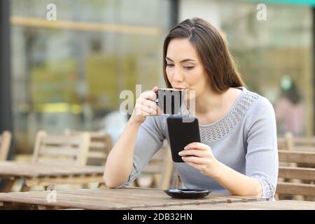 Giovane donna bevendo una tazza di caffè controllando il suo smartphone su una terrazza della caffetteria Foto Stock