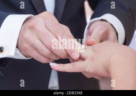 Marito con abito scuro e moglie con abito luminoso scambiano anelli di lusso dorati. Tenere le mani e mettere un anello su un dito l'altro. Foto Stock