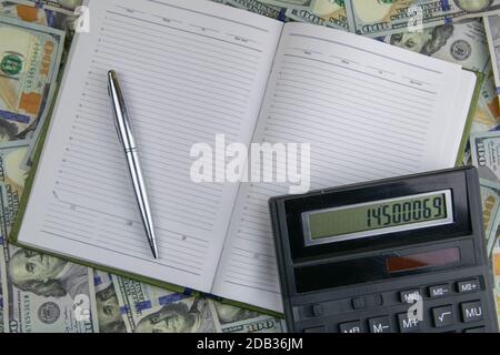 Un taccuino aperto con una penna e un supporto per calcolatrice su un mucchio di banconote da bambola. Foto Stock