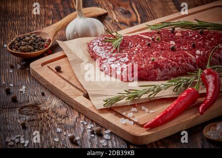 Carne fresca cruda. Bistecca di manzo su tagliere di legno chiaro, ramoscelli di rosmarino, spezie e verdure su sfondo di legno marrone. Menu bistecca Foto Stock