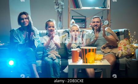 Saluto. Buona famiglia guardando proiettore, TV, film con pop corn e bevande la sera a casa. Madre, padre e bambini che trascorrono il tempo insieme. Comfort domestico, tecnologie moderne, concetto di emozioni. Foto Stock