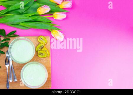 Yogurt al limone alla menta con kiwi in ciotole per insalata su tavola di legno insieme a tulipani su sfondo rosa. Foto Stock