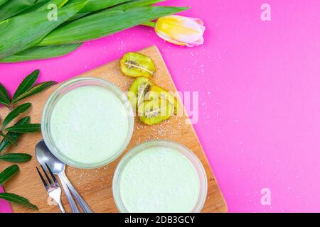 Yogurt al limone alla menta con kiwi in ciotole per insalata su tavola di legno insieme a tulipani su sfondo rosa. Foto Stock