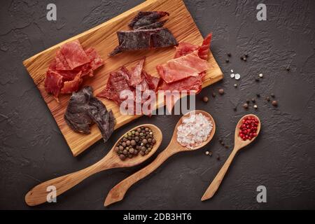 A scatti. Set di vari tipi di carne secca speziata su vassoio di legno, diversi peppercorn e sale su cucchiai di legno su fondo grigio scuro. Vista dall'alto. Snac Foto Stock