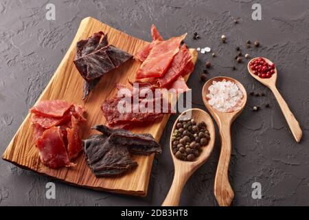 A scatti. Set di vari tipi di carne secca speziata su vassoio di legno, diversi peppercorn e sale su cucchiai di legno su fondo grigio scuro. Vista dall'alto. Snac Foto Stock