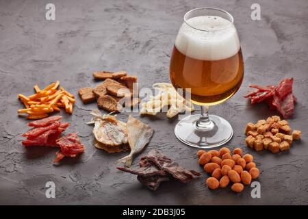 Bicchiere con birra e set di spuntini di vario tipo: Jerky, patatine, noci salate, pesce, cracker piccanti di pane, calamari secchi su sfondo grigio scuro Foto Stock