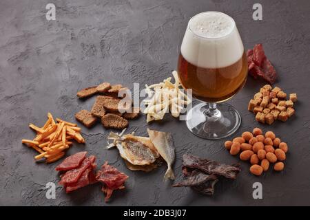 Bicchiere con birra e set di spuntini di vario tipo: Jerky, patatine, noci salate, pesce, cracker piccanti di pane, calamari secchi su sfondo grigio scuro Foto Stock