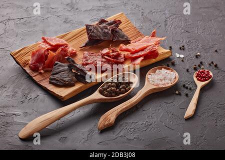 A scatti. Set di vari tipi di carne secca speziata su vassoio di legno, diversi peppercorn e sale su cucchiai di legno su fondo grigio scuro. Snack per la birra Foto Stock