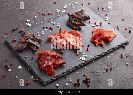 A scatti. Set di vari tipi di carne secca speziata su tagliere in pietra nera su fondo grigio scuro. Snack per la birra. Foto Stock