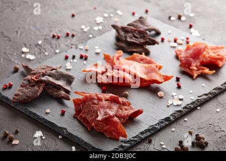 A scatti. Set di vari tipi di carne secca speziata su tagliere in pietra nera su fondo grigio scuro. Snack per la birra. Foto Stock