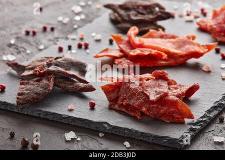 A scatti. Set di vari tipi di carne secca speziata su tagliere in pietra nera su fondo grigio scuro. Snack per la birra. Foto Stock