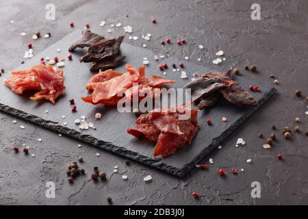 A scatti. Set di vari tipi di carne secca speziata su tagliere in pietra nera su fondo grigio scuro. Snack per la birra. Foto Stock