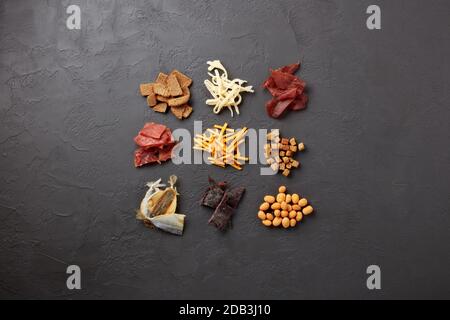 Set di vari tipi di snack: Jerky, patatine, noci salate, pesce di mare, cracker piccanti di pane, calamari secchi su fondo grigio scuro. Vista dall'alto. Snac Foto Stock