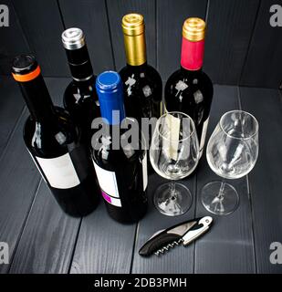 Bottiglie di vino rosso di produttori diversi con due bicchieri di vino frizzante e un cavatappi per la degustazione, in uno spazio di legno grigio ben illuminato Foto Stock