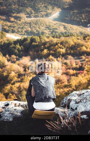 Ragazza seduta meditando su una cima di montagna. Distanza sociale in escursioni da soli lontano dai luoghi sociali. Vista dall'alto Foto Stock