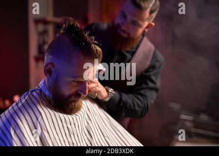 il sicuro hairstylist maschile tagliare i capelli a un elegante uomo seduto in salone, giovane maschio godere del processo di taglio Foto Stock