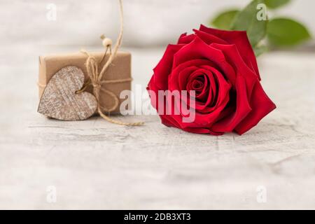 Regalo avvolto in carta artigianale accanto alla rosa rossa su sfondo di pietra bianca. Regalo romantico per la giornata di San Valentino con spazio per il testo. Concetto di proposta di matrimonio. Messa a fuoco morbida. Spazio di copia Foto Stock