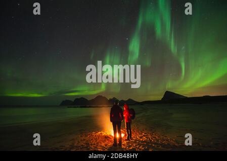 Due persone si riscaldano da un falò sulla spiaggia con luci a nord - aurora borealis nel cielo sopra, Flakstadøy, Isole Lofoten, Norvegia Foto Stock