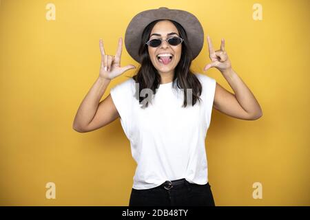 Bella donna che indossa occhiali da sole, casual t-shirt bianca e un cappello in piedi su sfondo giallo gridando con pazzo espressione facendo rock simbolo wi Foto Stock