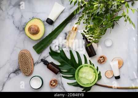 Cosmetici naturali con ingredienti a base di erbe, piatto. Bottiglie di olii essenziali e un vaso di idratante e foglie verdi su sfondo di marmo di pietra bianca e concetto di stile di vita sano. Foto Stock