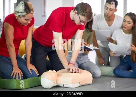Giovane maschio che pratica CPR primo soccorso su un manichino in presenza di persone, al chiuso Foto Stock