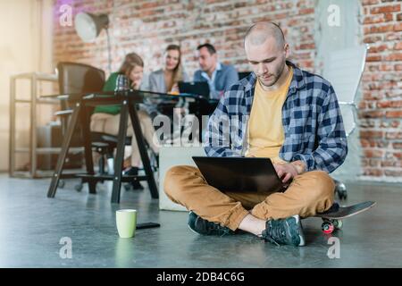 Assistente di anca in una codifica di avvio con un laptop seduto sullo skateboard con i colleghi in background Foto Stock