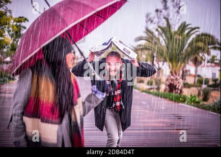 La coppia amorevole sotto la pioggia - bella ragazza che tiene ombrello mentre il suo ragazzo corre a lei sotto la pioggia Sotto un giornale - sta girando e. Foto Stock