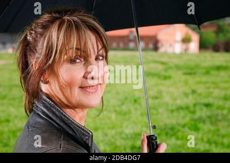 Vista laterale testa e spalle di una donna di mezza età che tiene un ombrello contro uno sfondo rurale sfocato sorridendo alla telecamera. Foto Stock