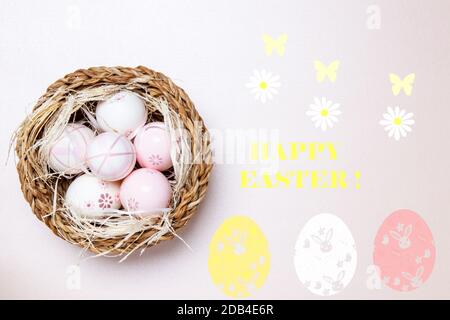 Buona cartolina di auguri di Pasqua. Vista dall'alto delle uova colorate con paglia in un cesto di vimini su uno sfondo rosa chiaro con iscrizione Happy easter e Bloss Foto Stock