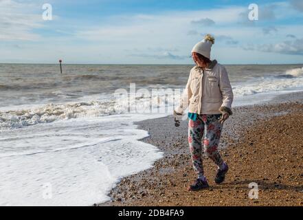 Donna anziana vestita di cappotto, cappello, guanti e pantaloni, camminando su una spiaggia vicino al mare in autunno nel Regno Unito. Passeggiata sul mare. Foto Stock