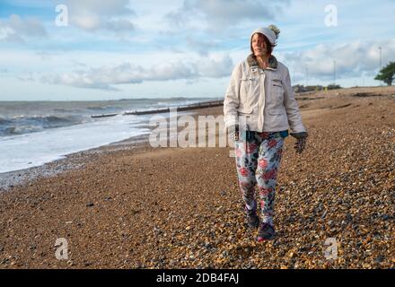 Donna anziana vestita di cappotto, cappello, guanti e pantaloni, camminando su una spiaggia vicino al mare in autunno nel Regno Unito. Passeggiata sul mare. Foto Stock