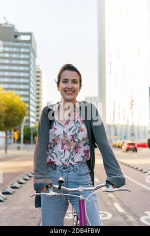 ritratto verticale di una giovane donna sorridente in città che cavalcava una bicicletta rosa retrò sulla pista ciclabile, concetto di stile di vita attivo e mobil sostenibile Foto Stock