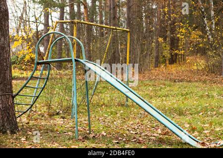 Parco giochi abbandonato in autunno. Un vecchio gol calcistico con una rete strappata e uno scivolo per bambini. Foto Stock