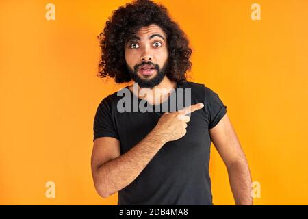 giovane arabo alato maschio con trendy punti hairdo con indice dito da parte, mostra qualcosa, ragazzo ricciolo in abbigliamento casual isolato su sfondo arancione Foto Stock