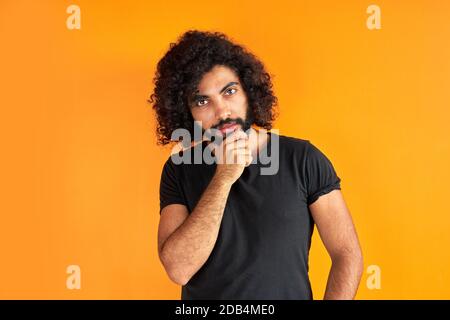 il maschio arabico determinato con la barba sta pensando e guardando preoccupato, in piedi contro sfondo arancione. Foto Stock