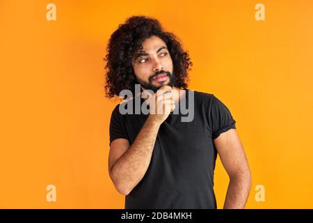 il maschio arabico determinato con la barba sta pensando e guardando preoccupato, in piedi contro sfondo arancione. Foto Stock