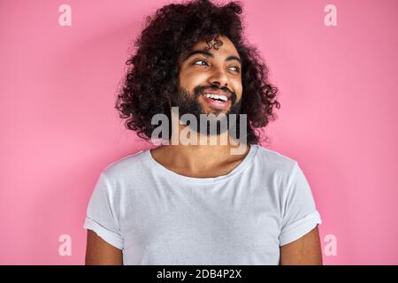ritratto di allegro positivo maschio di aspetto arabico guardando via ridendo, ascolta scherzo, divertirsi con qualcuno, isolato su sfondo rosa Foto Stock