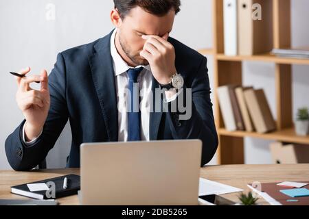 Uomo d'affari stanco con emicrania, tenendo la mano vicino agli occhi, mentre si siede sul posto di lavoro su sfondo offuscato Foto Stock