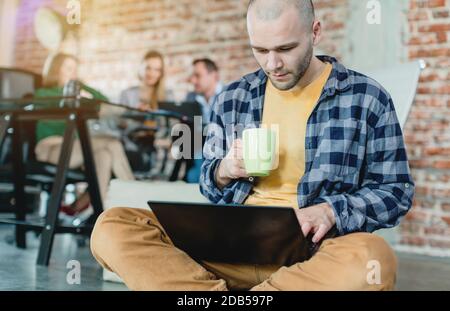 Assistente di anca in una codifica di avvio con un laptop seduto sullo skateboard con i colleghi in background Foto Stock