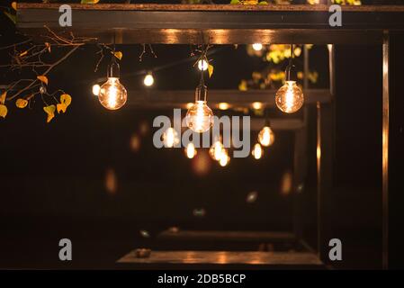 Molte lampadine elettriche a incandescenza di notte sopra il tavoli tra gli alberi del parco cittadino Foto Stock