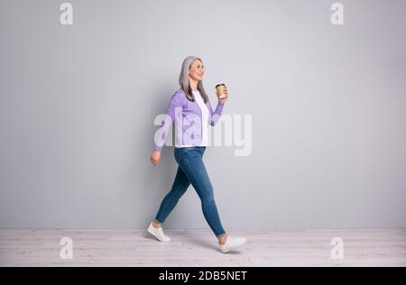 Foto a lunghezza intera del corpo di una donna anziana sorridente che ha rompere andando veloce mantenere la tazza di caffè indossando jeans cardigan occhiali isolati su grigio Foto Stock